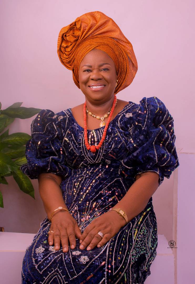 Mrs. Ebunoluwa Annah Oladele (nee Akeremale) smiling, wearing an orange gele, navy dress and coral beads.